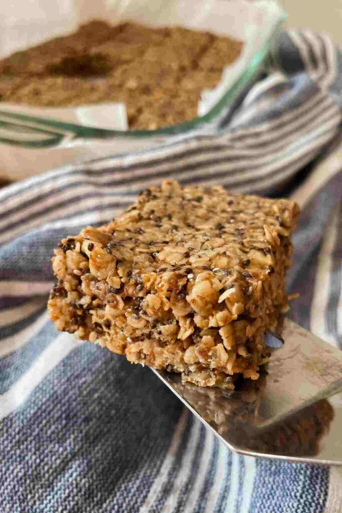 Homemade chewy granola bar on serving utensil in front of blue striped kitchen towel and baking pan of granola bars