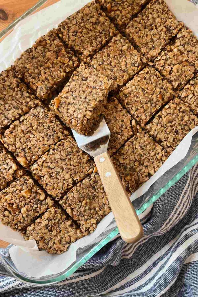 Homemade chewy granola bars in pan lined with parchment paper, one granola bar being lifted out by server with wooden handle
