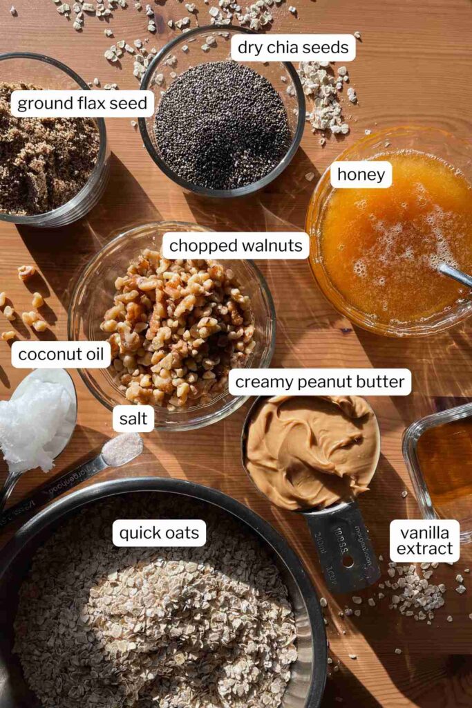 Overhead shot of easy chewy granola bar recipe ingredients in bowls and labeled: dry chia seeds, ground flax seed, chopped walnuts, honey, coconut oil, salt, creamy peanut butter, vanilla extract, quick oats