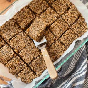 Homemade chewy granola bars in pan lined with parchment paper, one granola bar being lifted out by server with wooden handle