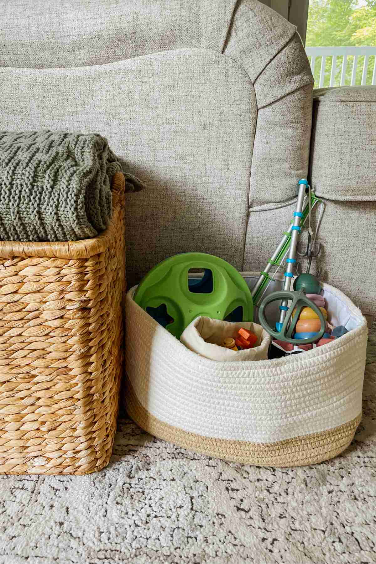 White basket filled with toys sitting next to a tan wicker basket with a green blanket visible on top next to a cream colored couch