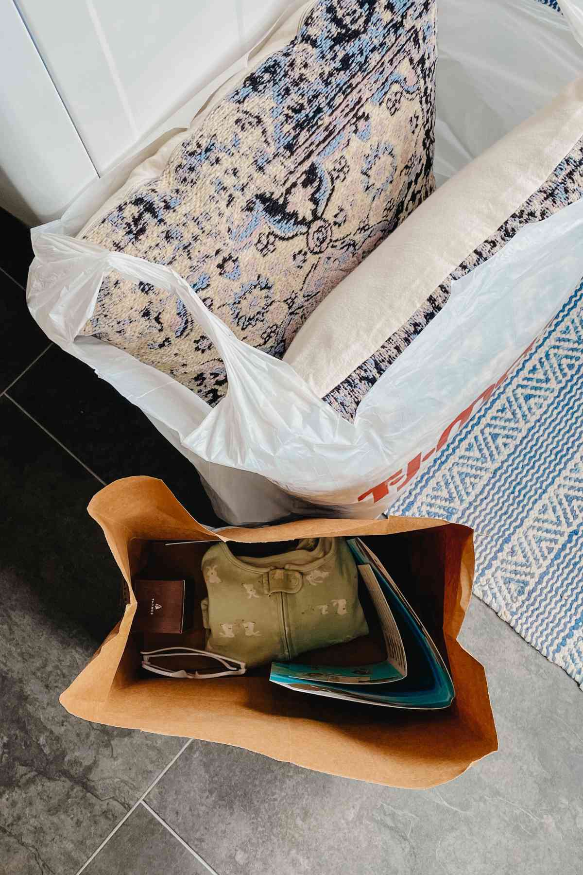 Plastic bag holding 2 decorative pillows and a paper bag holding toys and clothes sitting on a grey laminate floor with a blue and cream striped rug underneath
