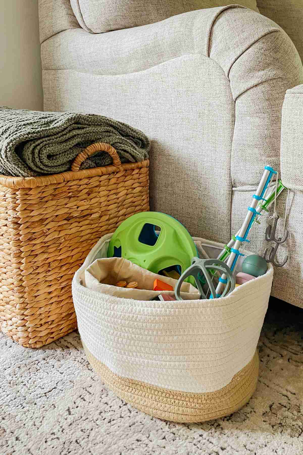 White basket filled with toys sitting next to a tan wicker basket with a green blanket visible on top next to a cream colored couch