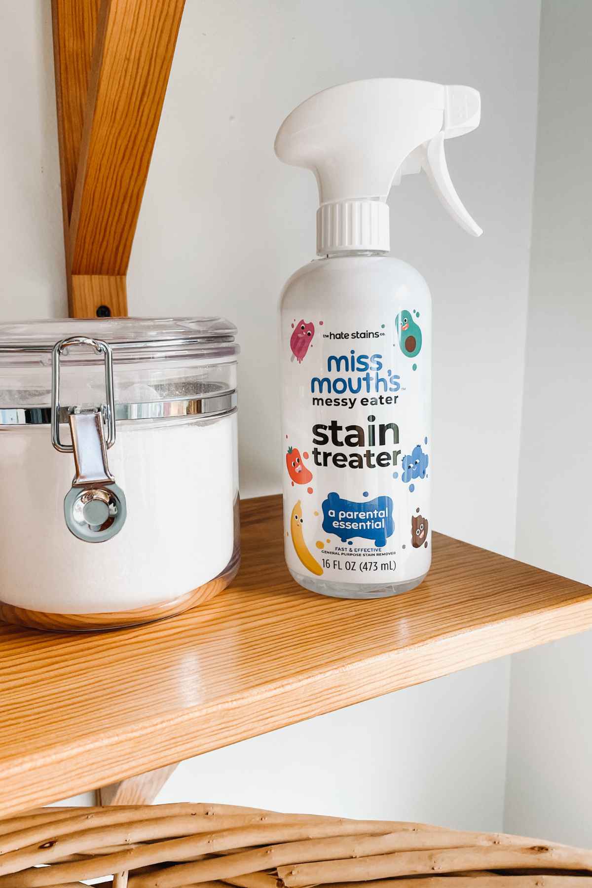 Bottle of Miss Mouth's messy eater stain remover and clear plastic canister of laundry detergent sitting on a wooden shelf with the edge of a wicker laundry basket visible below