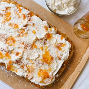 Peaches and cream sweet sourdough focaccia sitting on parchment paper on a wooden cutting board with a jar of peach preserves and homemade whipped cream in a glass bowl beside