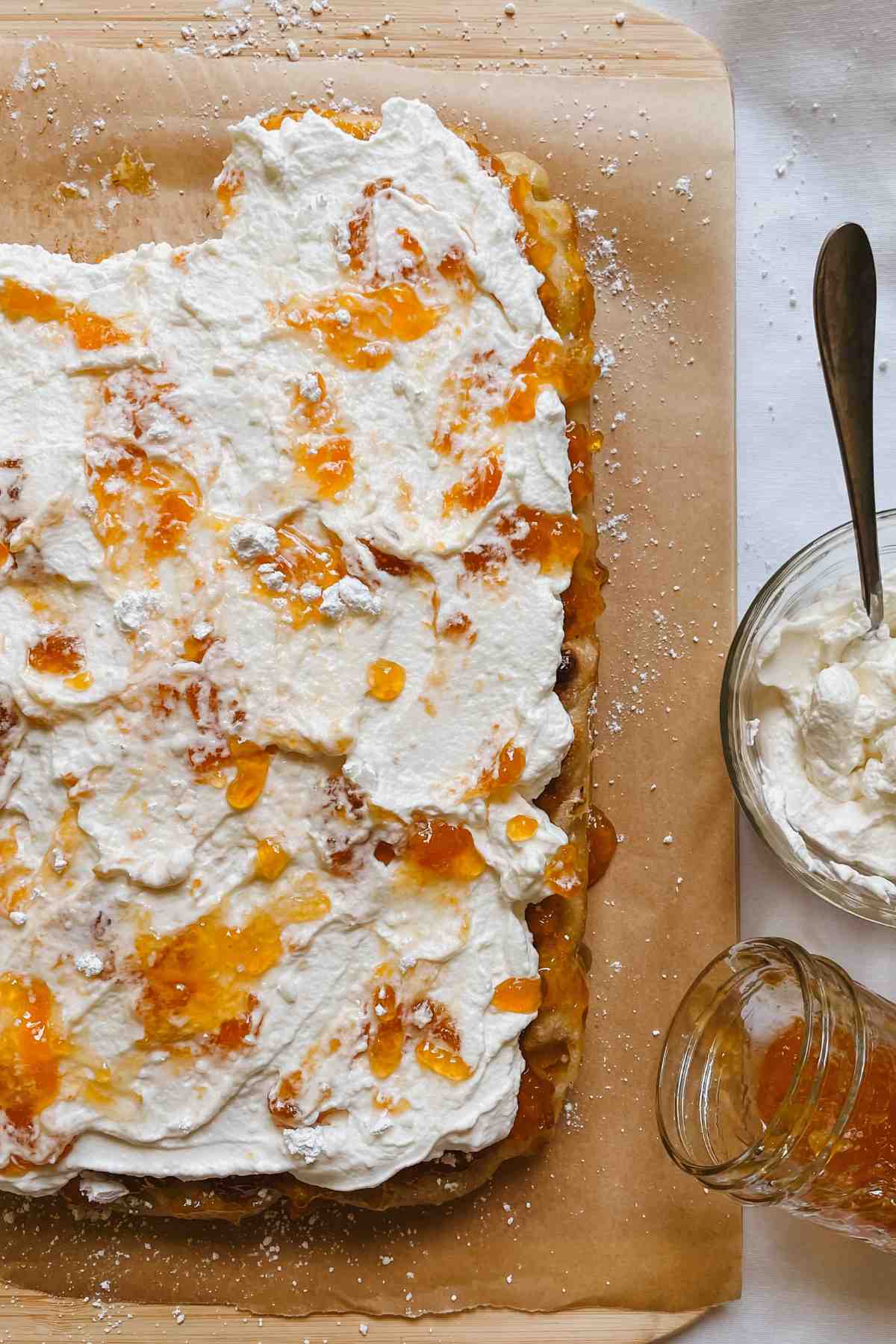 Peaches and cream sweet sourdough focaccia sitting on parchment paper on a wooden cutting board with a jar of peach preserves and homemade whipped cream in a glass bowl beside