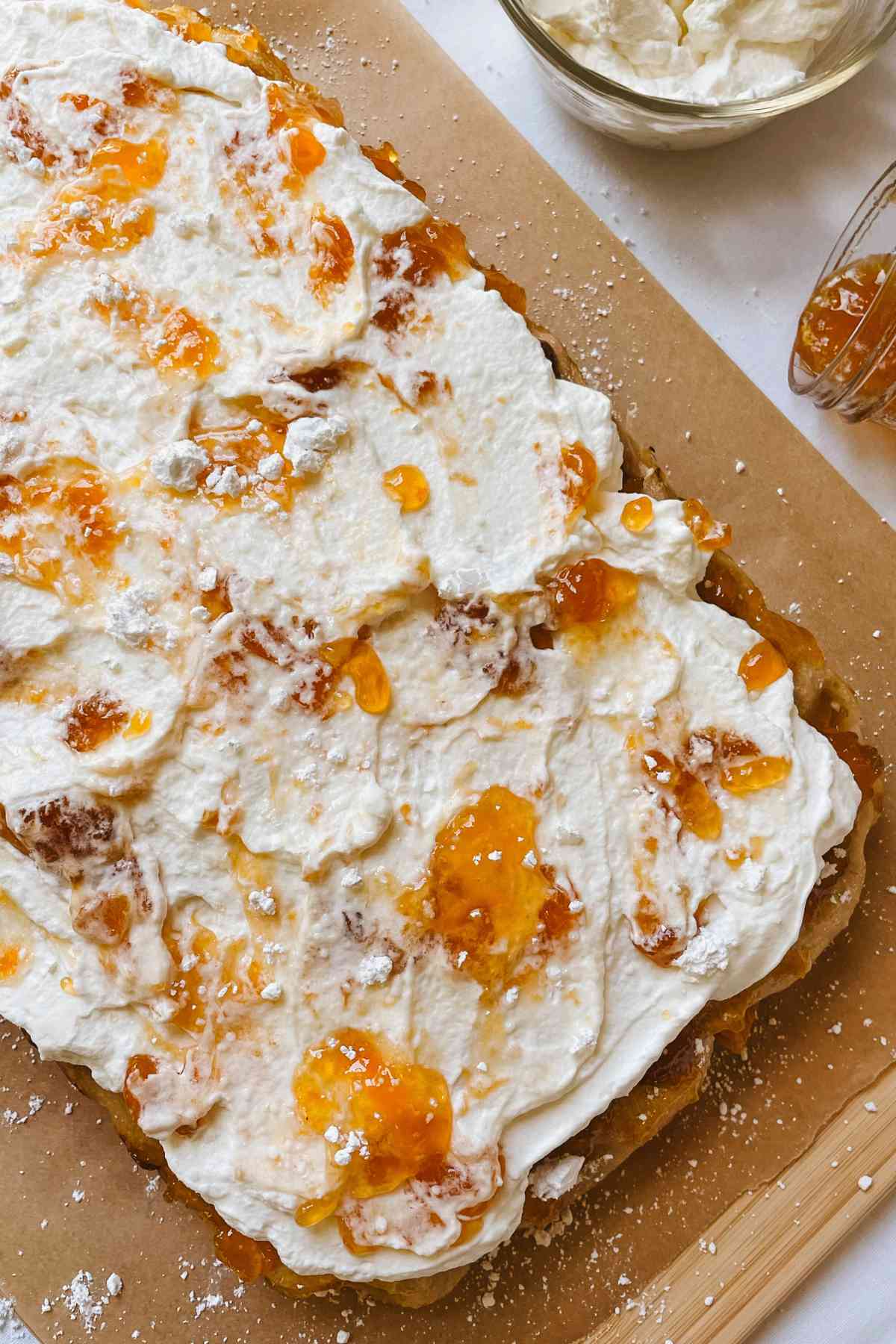 Peaches and cream sweet sourdough focaccia topped with homemade whipped cream and peach preserves, sitting on parchment paper on a wooden cutting board with a jar of peach preserves and homemade whipped cream in a glass bowl beside