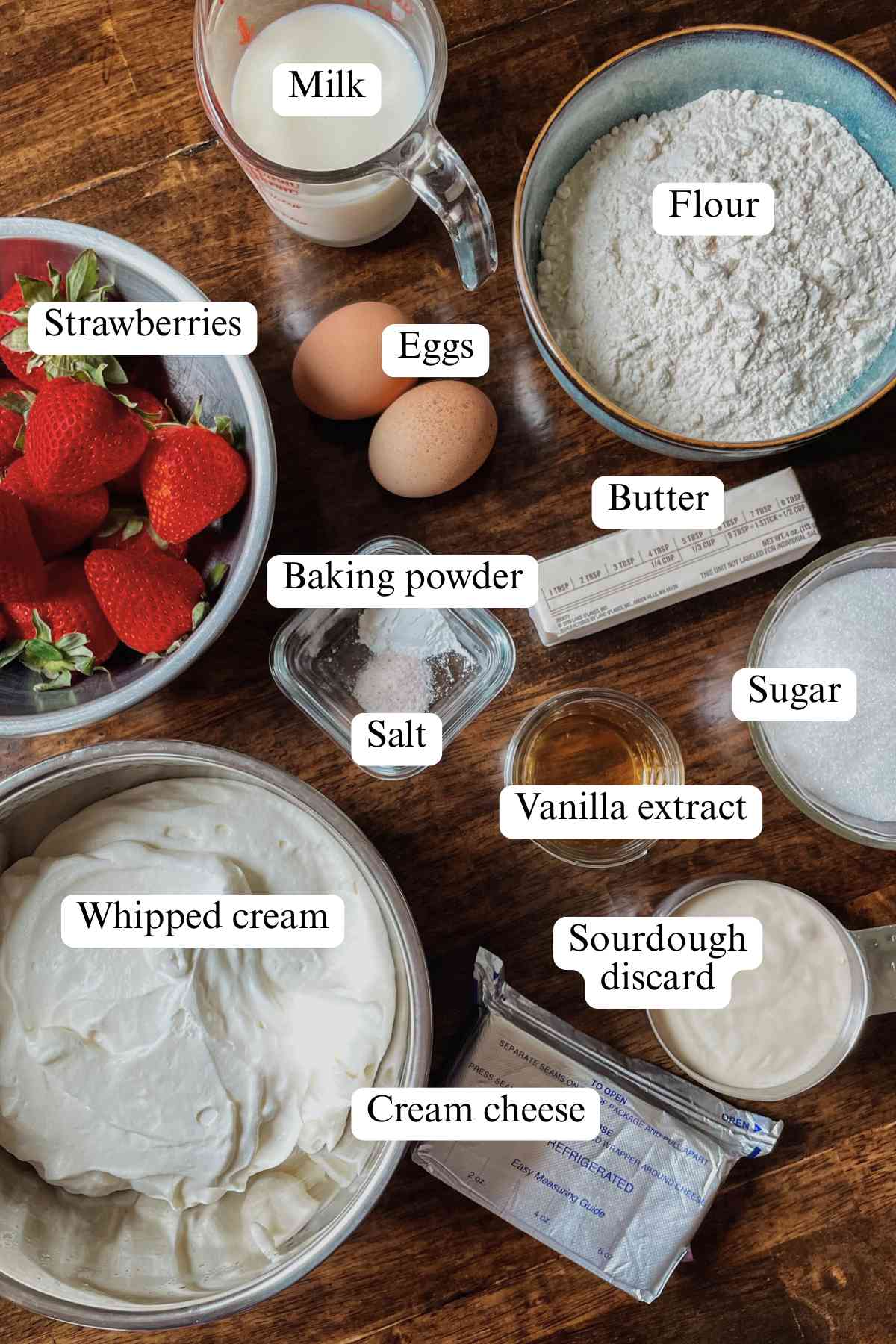 Sourdough discard strawberry shortcake bars ingredients sitting on table in bowls labeled: milk, strawberries, eggs, flour, butter, baking powder, salt, sugar, vanilla extract, sourdough discard, whipped cream, cream cheese