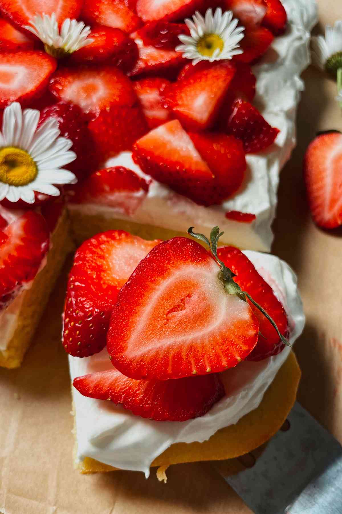 Slice of sourdough discard strawberry shortcake bar with layer of shortcake, cream cheese frosting, and topped with sliced strawberries sitting on brown parchment paper with sourdough strawberry shortcake in the background