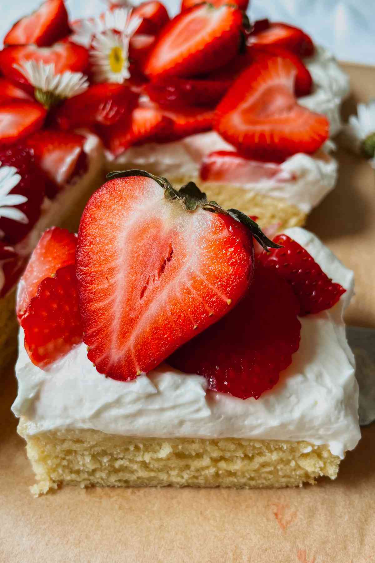 Slice of sourdough discard strawberry shortcake bar with layer of shortcake, cream cheese frosting, and topped with sliced strawberries sitting on brown parchment paper with sourdough strawberry shortcake in the background