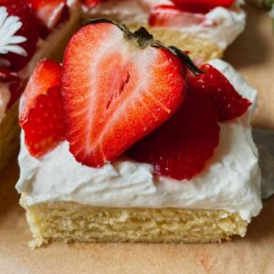 Slice of sourdough discard strawberry shortcake bar with layer of shortcake, cream cheese frosting, and topped with sliced strawberries sitting on brown parchment paper with sourdough strawberry shortcake in the background