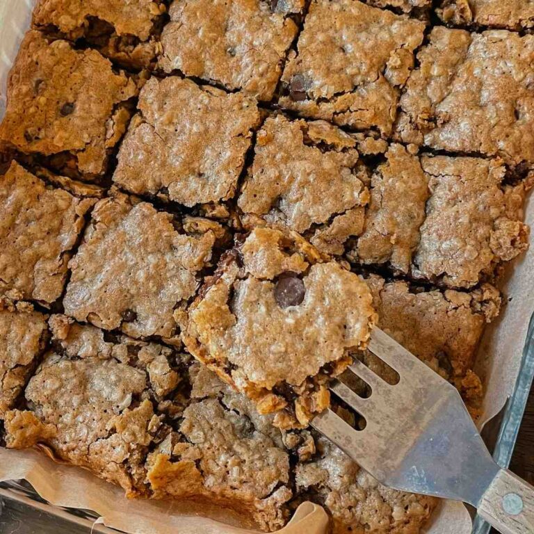 Sliced squares of sourdough oatmeal chocolate chip cookie bars in a parchment lined baking pan with a serving utensil holding a bar above the rest
