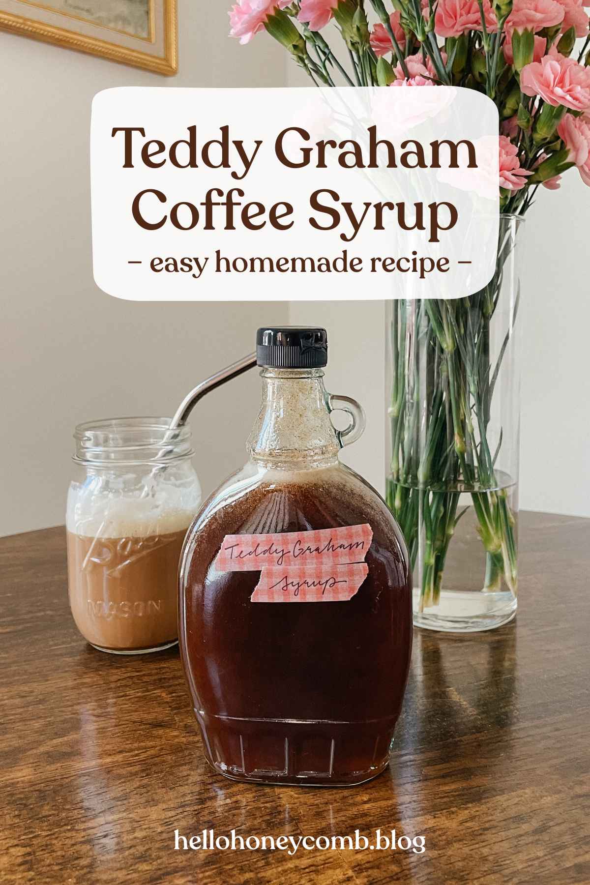 Glass bottle of teddy graham latte coffee syrup sitting on wooden table with iced latte and vase of pink flowers in background with text overlaid that reads Teddy Graham Coffee Syrup easy homemade recipe