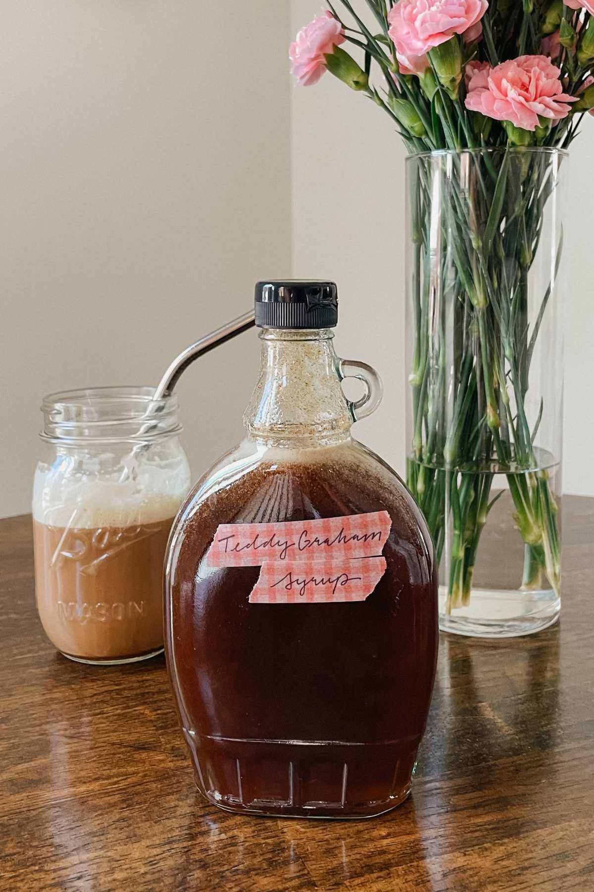 Glass bottle of teddy graham latte coffee syrup sitting on wooden table with iced latte and vase of pink flowers in background