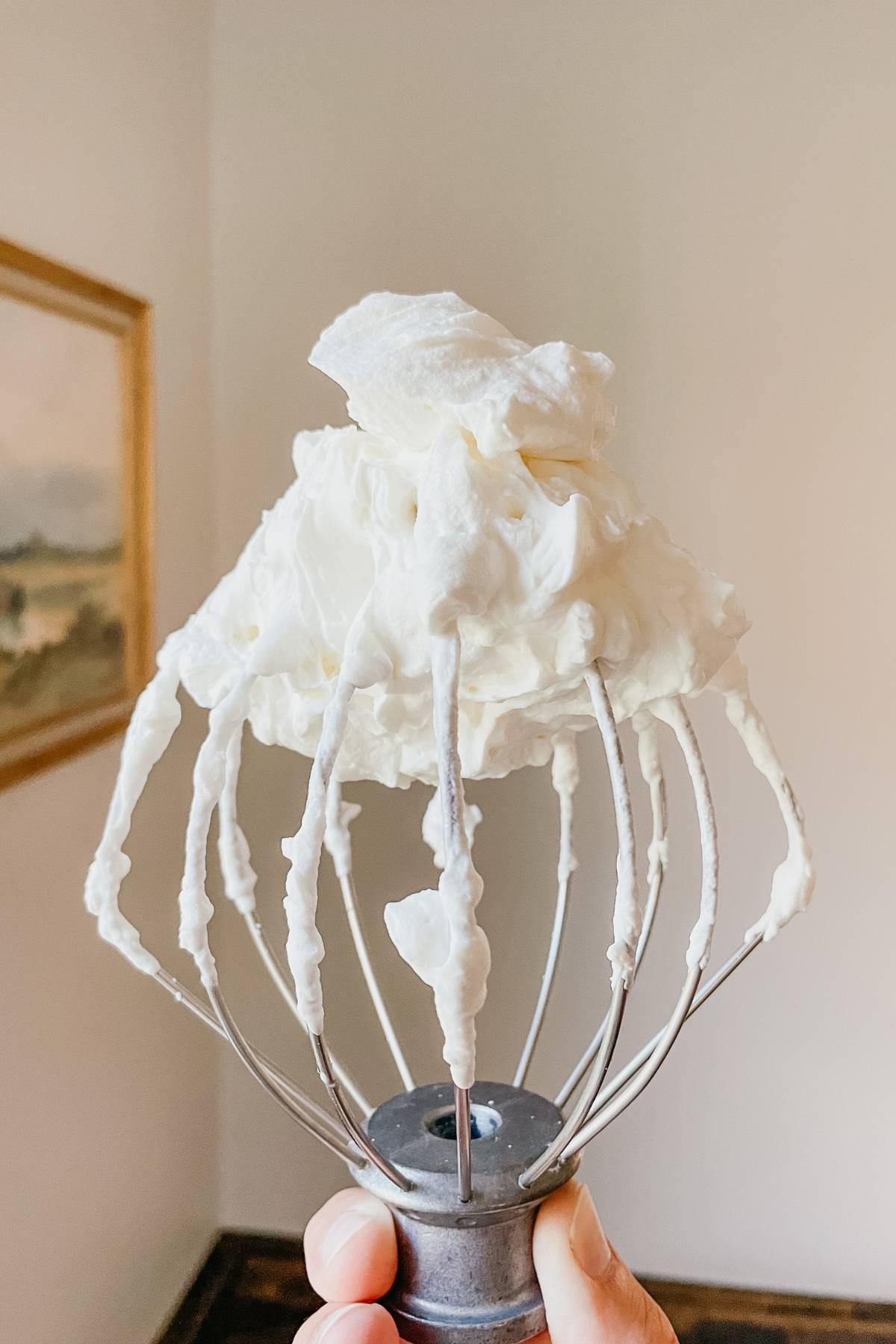 Hand holding a wire whisk with homemade whipped cream in front of a wall