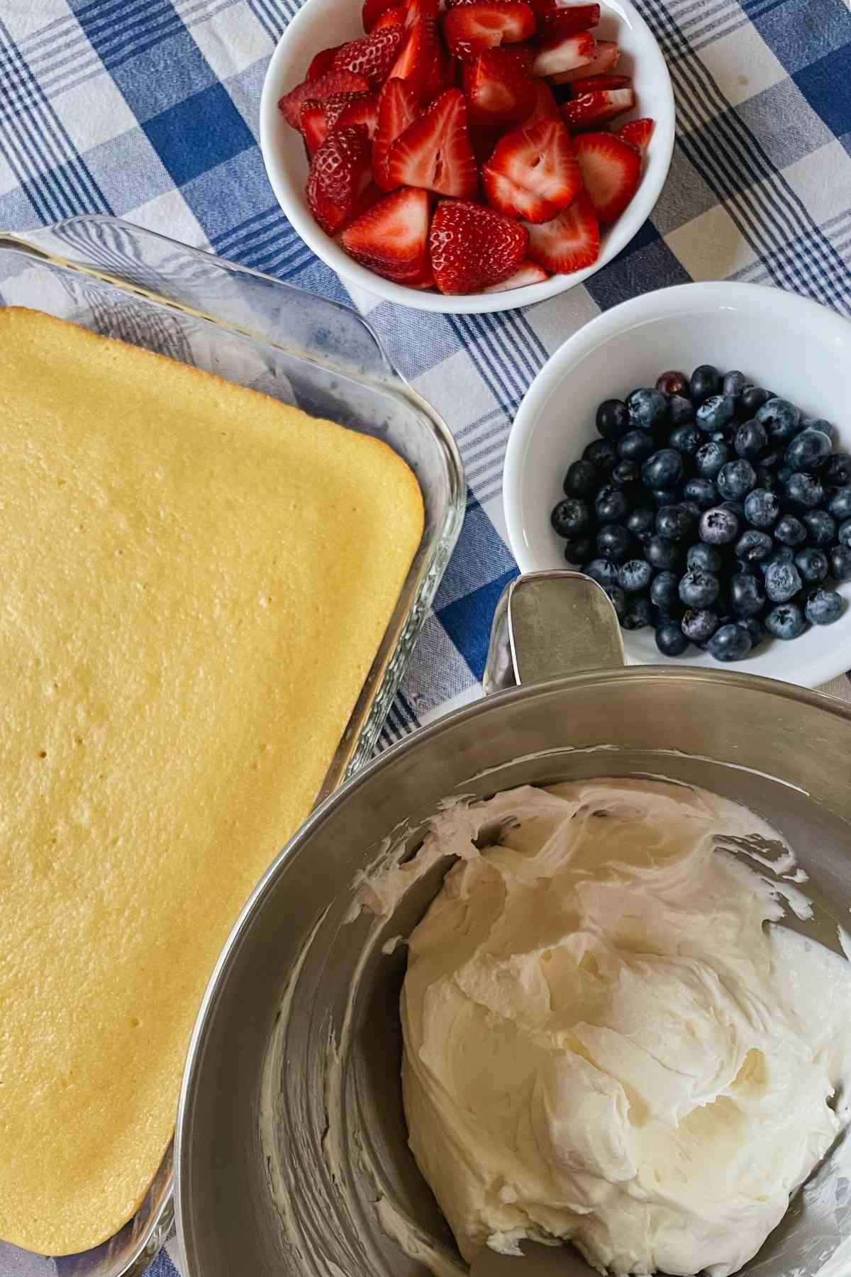Sourdough shortcake in a pan, blueberries and strawberries in bowls, and cream cheese icing in mixing bowl on a blue checkered tablecloth