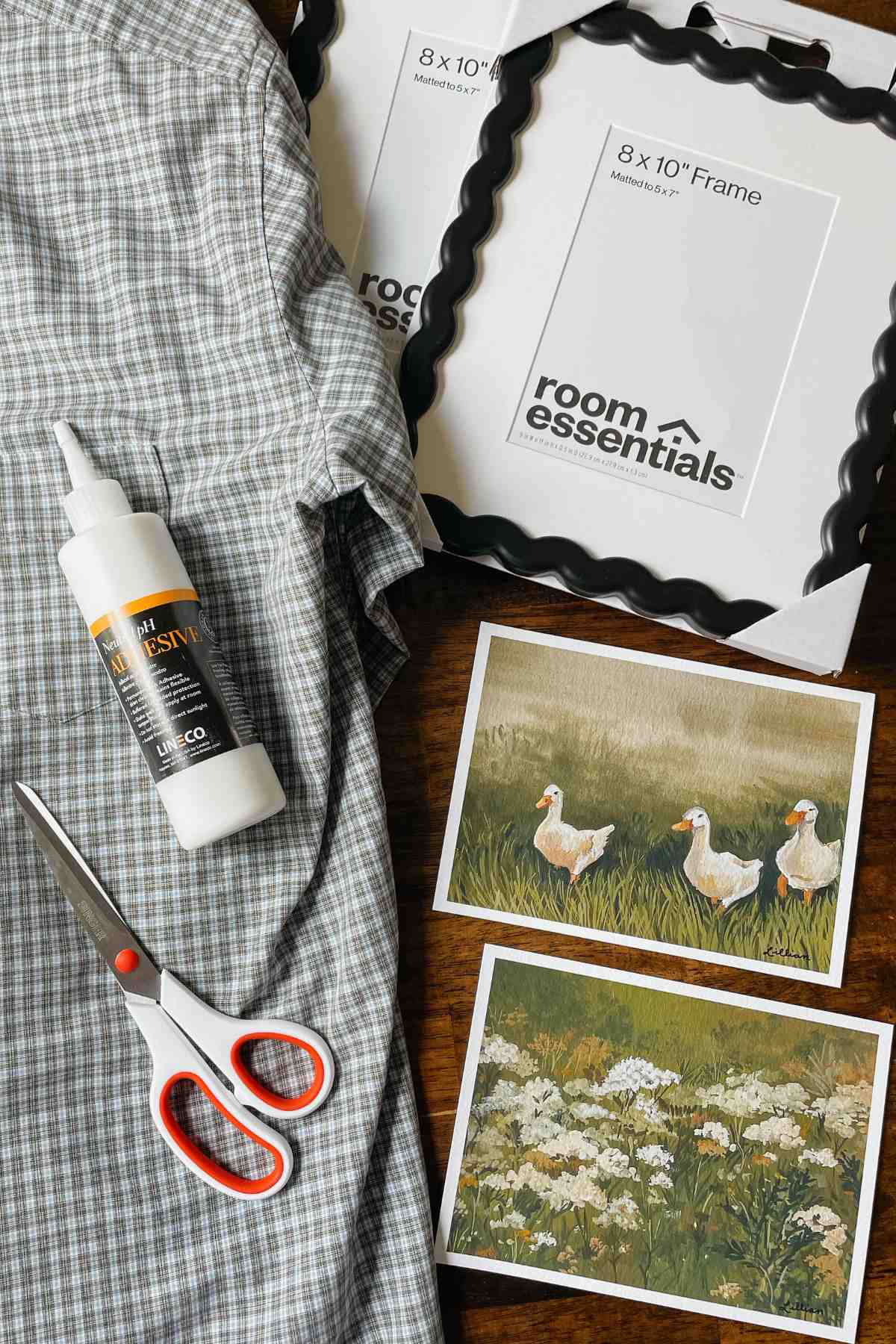 Black wavy picture frames, plaid men's shirt, scissors, bottle of glue, and art prints laid on a wooden table