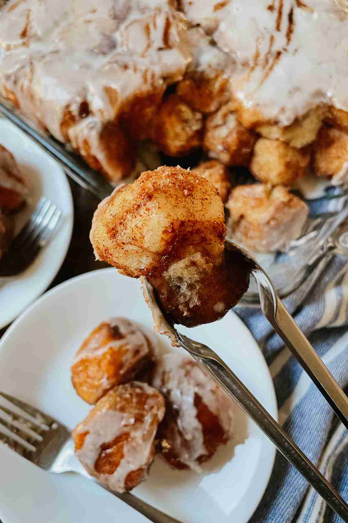 Piece of sourdough brown butter monkey bread held by tongs in front of a pan of monkey bread and white plates with forks