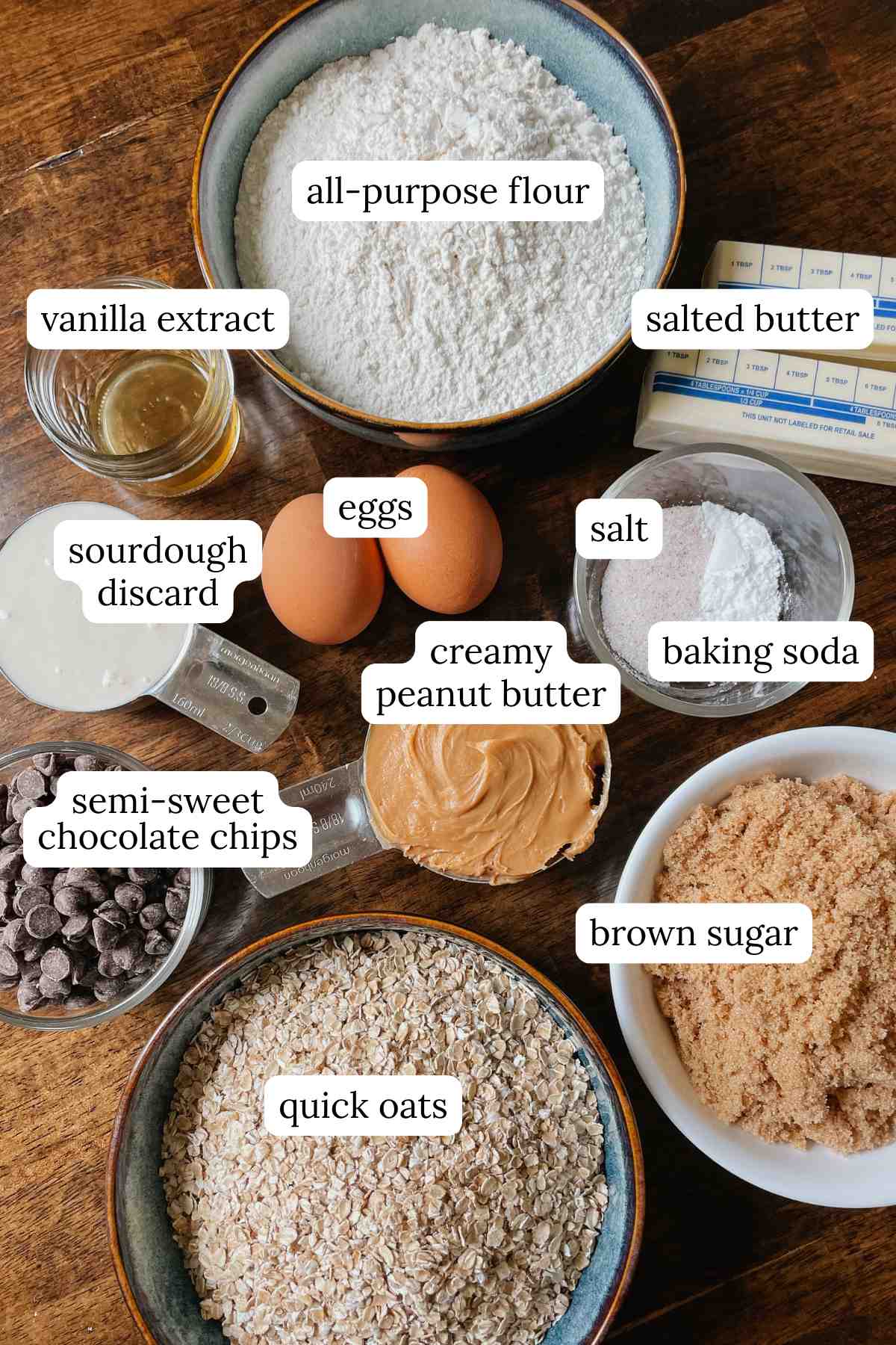 Ingredients for sourdough peanut butter oatmeal chocolate chip cookie bars in bowls labeled on wooden table