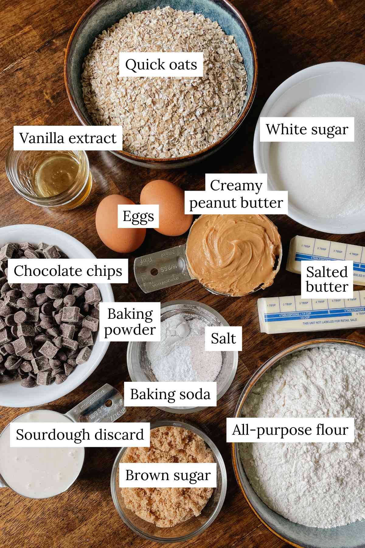 Ingredients for sourdough peanut butter oatmeal chocolate chip cookies in bowls labeled on wooden table