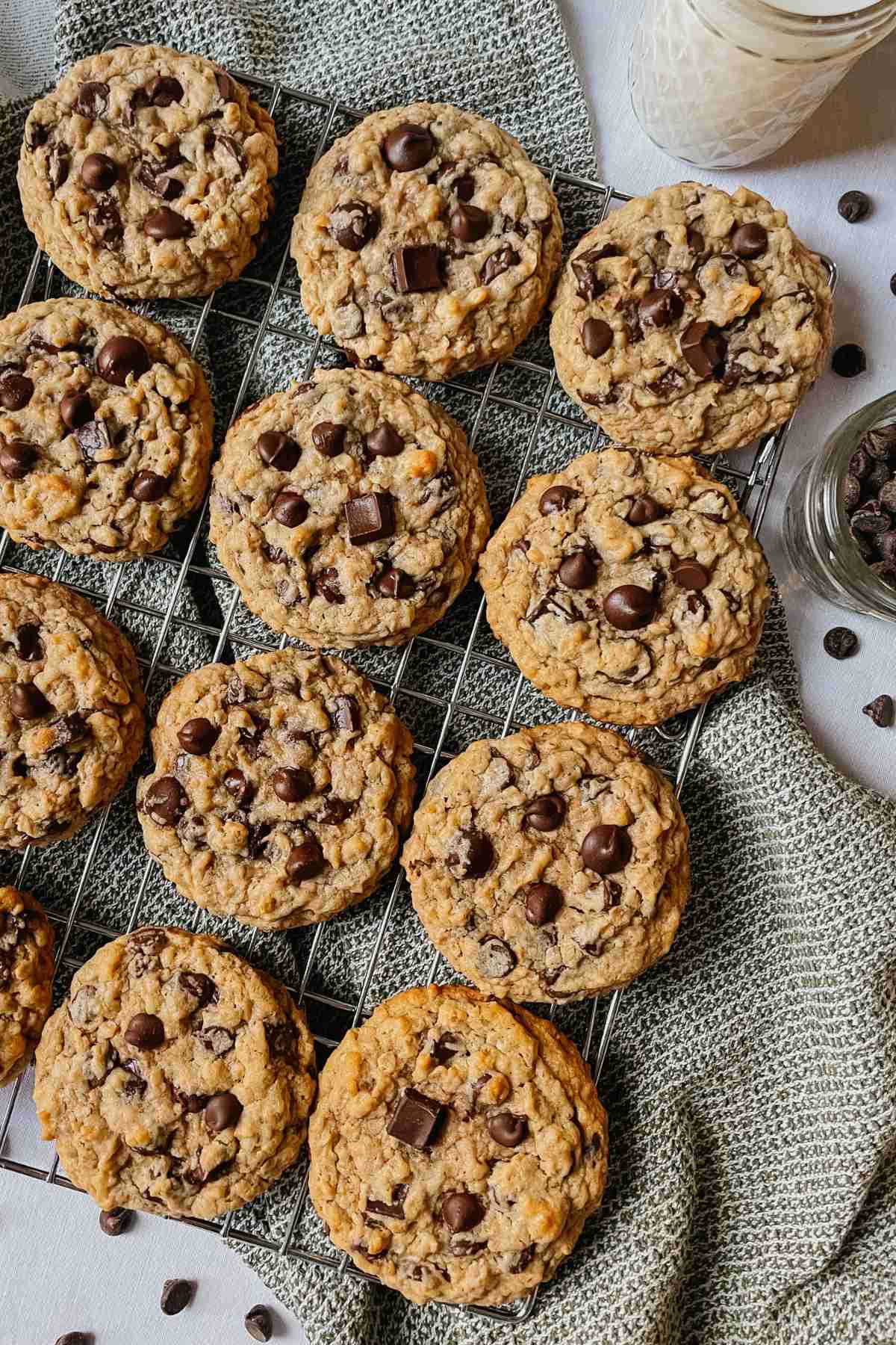 Sourdough peanut butter oatmeal chocolate chip cookies on wire rack with green kitchen towel, chocolate chips, and glass of milk around