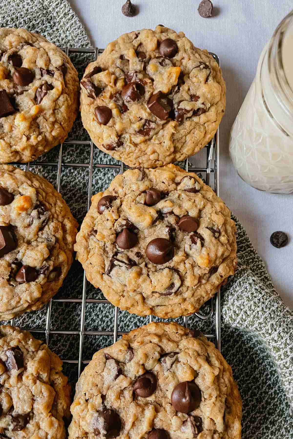 Sourdough peanut butter oatmeal chocolate chip cookies on wire rack with green kitchen towel, chocolate chips, and glass of milk around