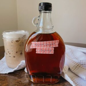 Glass bottle of brown sugar coffee syrup sitting on wooden table with iced coffee and kitchen towel behind