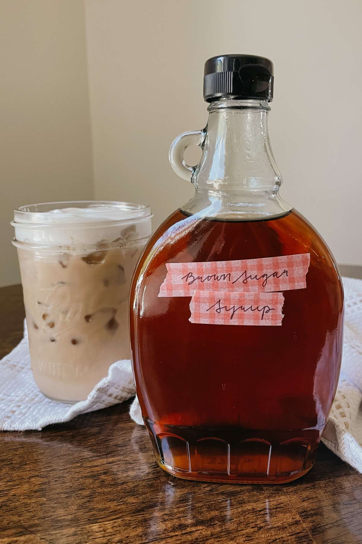 Glass bottle of brown sugar coffee syrup sitting on wooden table with iced coffee and kitchen towel behind