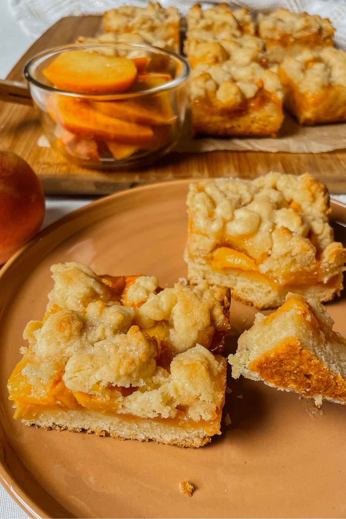 Sourdough peach crumb bar squares on orange plate with peaches and crumb bars in the background
